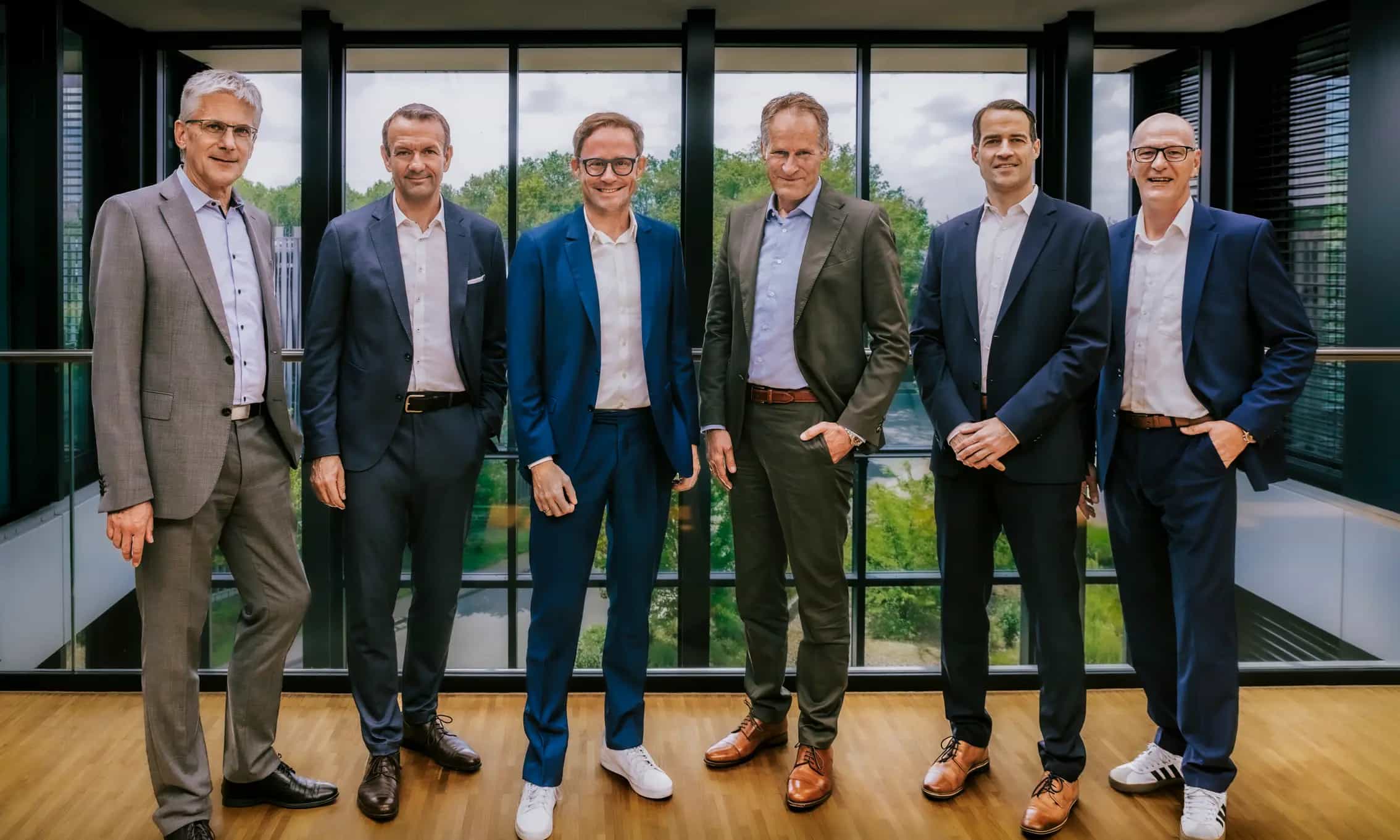 Six executives in business suits are standing in a modern office lounge in front of a large set of windows.