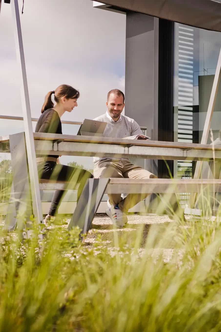 Two people work on laptops on a modern terrace with plants in the foreground.