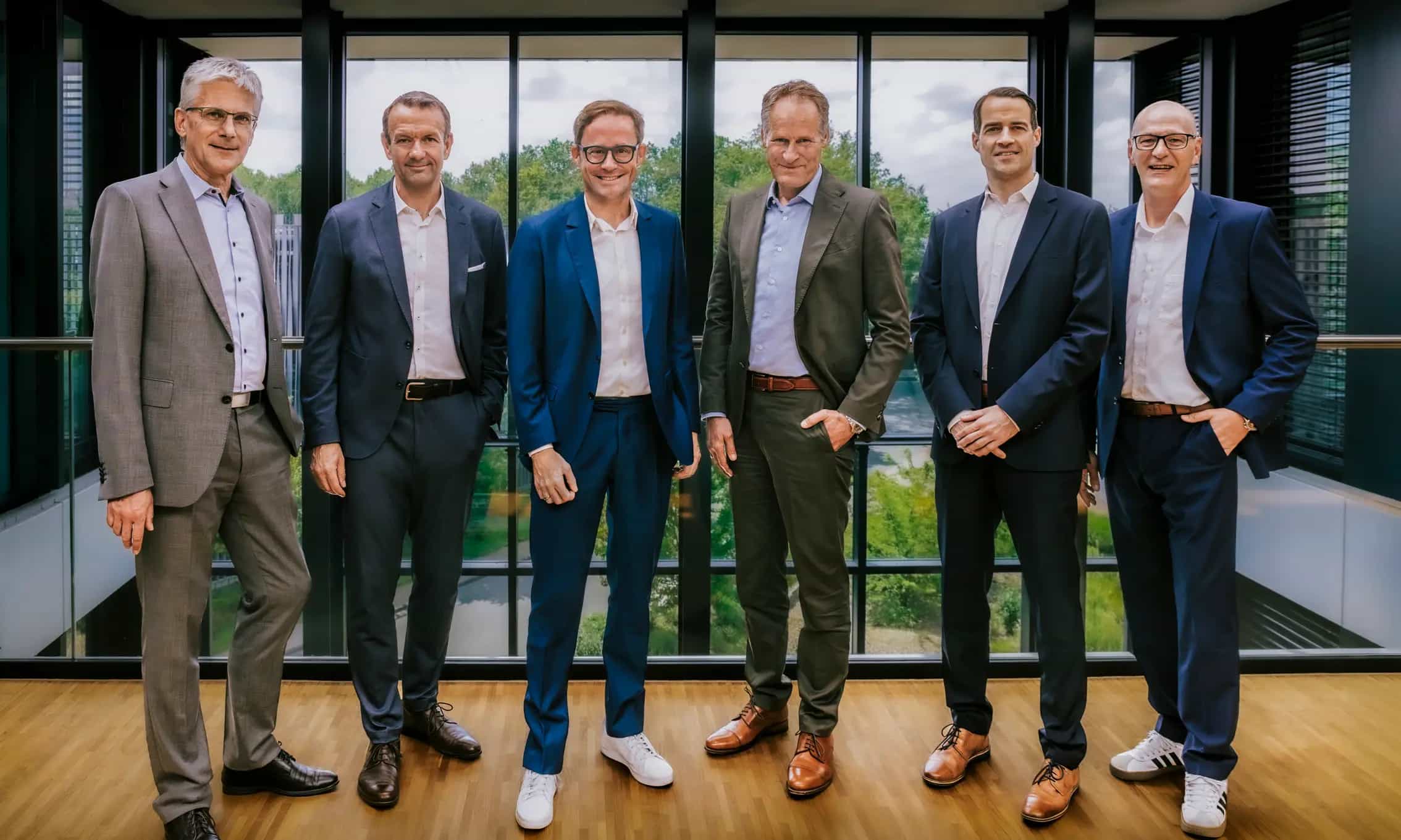 Gruppe von sechs Geschäftsführern in Anzügen in einem modernen Büro mit großen Fenstern im Hintergrund.