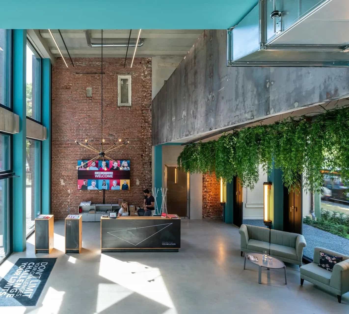 Modern hotel lobby area with a high ceiling, blue walls, a reception desk, green plants on the ceiling and an inviting seating area.